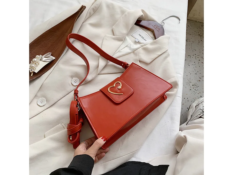 A red small square shoulder bag with a gold heart detail held by a hand.