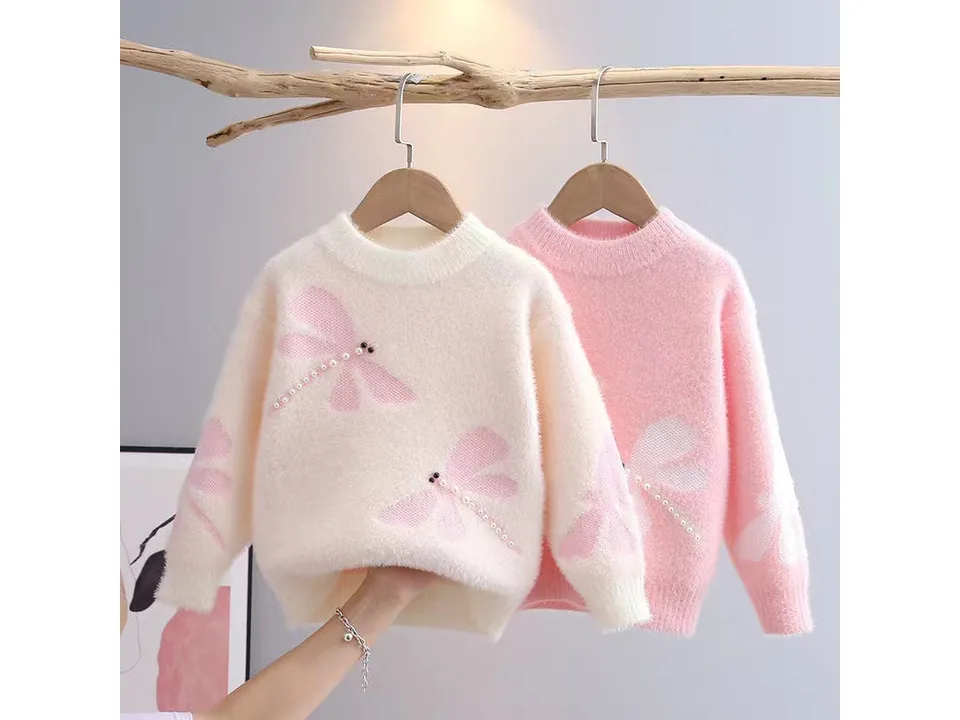 Two fuzzy children's sweaters, one cream and one pink, both with dragonfly patterns, hanging on a wooden branch.