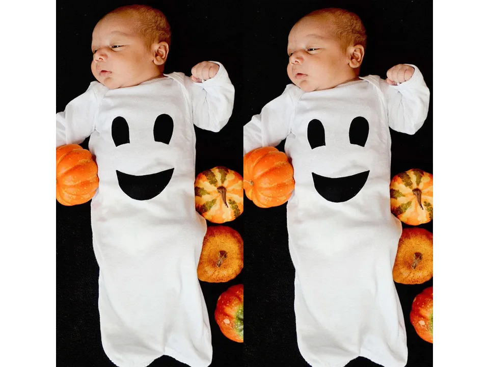 A baby wearing a white long-sleeve ghost-face onesie lying next to small pumpkins.