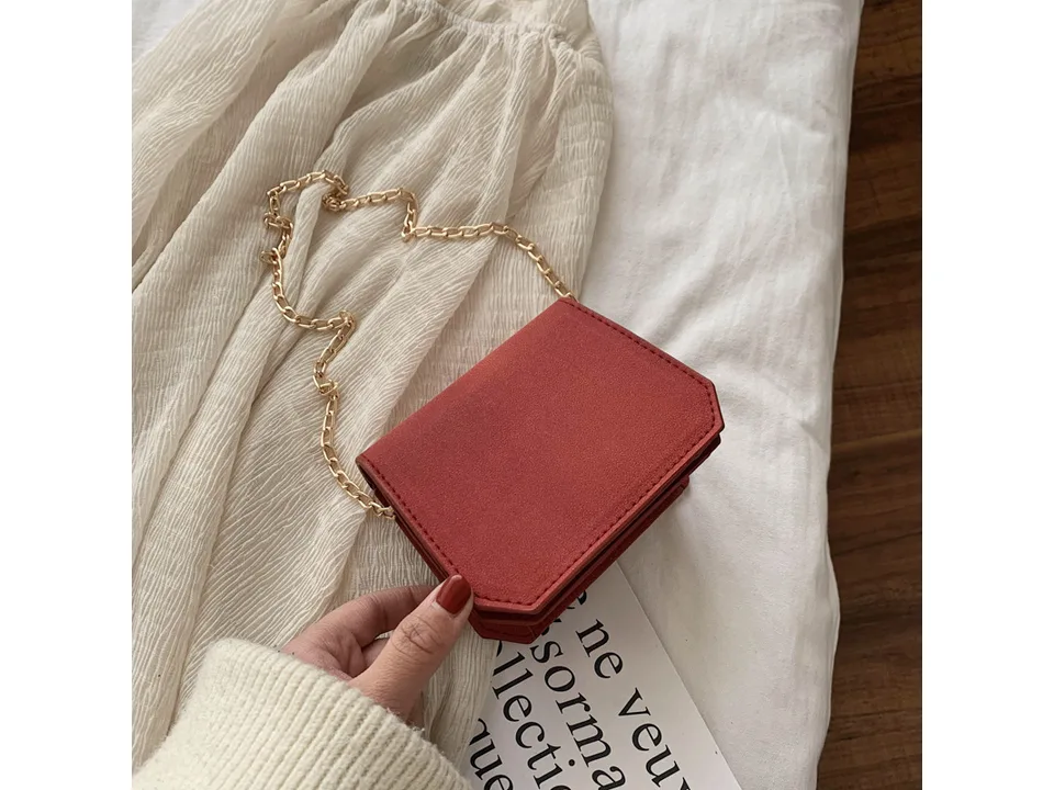 A small red suede-like shoulder bag with a gold chain strap held in a hand.