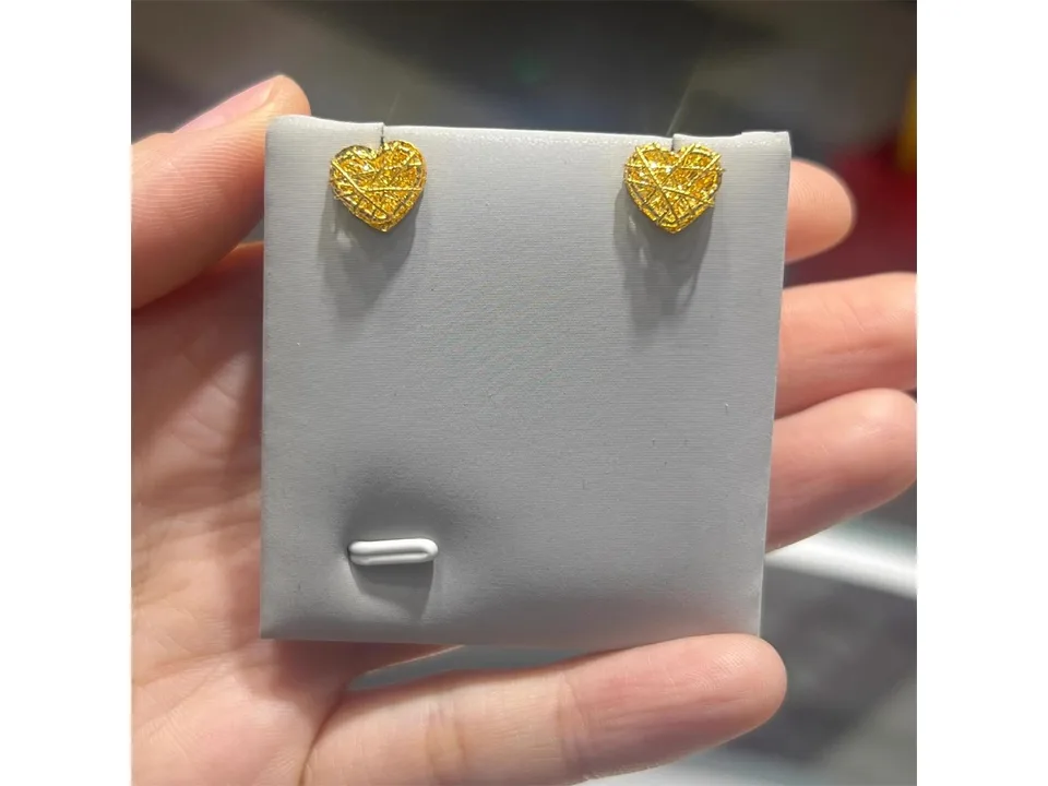 A pair of gold heart stud earrings on a grey display card held in a hand.
