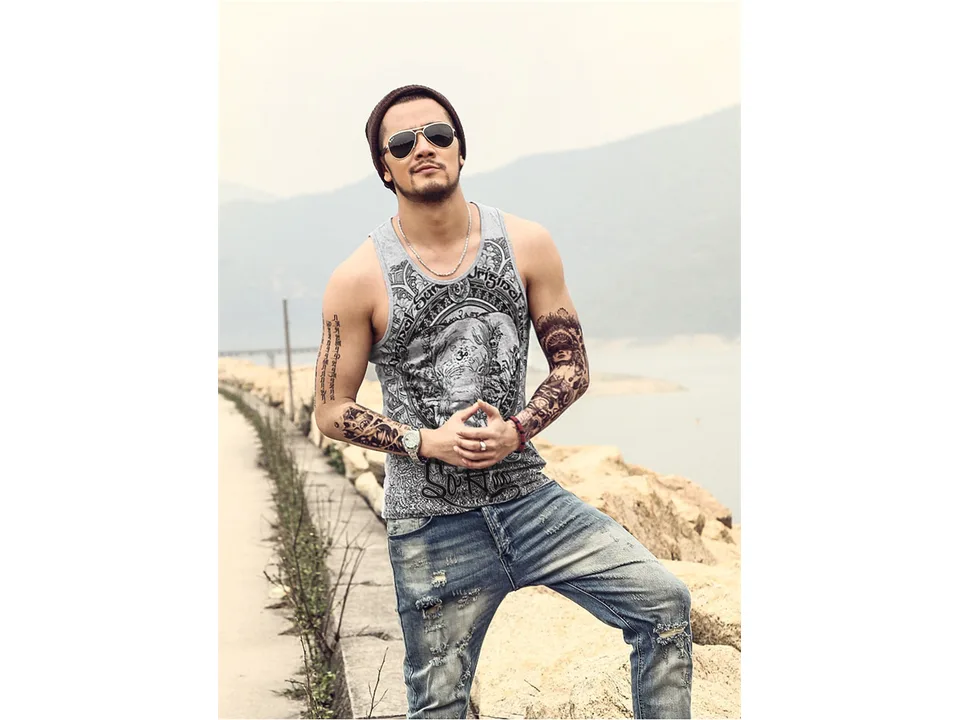 A man wearing a grey tank top with an elephant print design, posing with hands together.