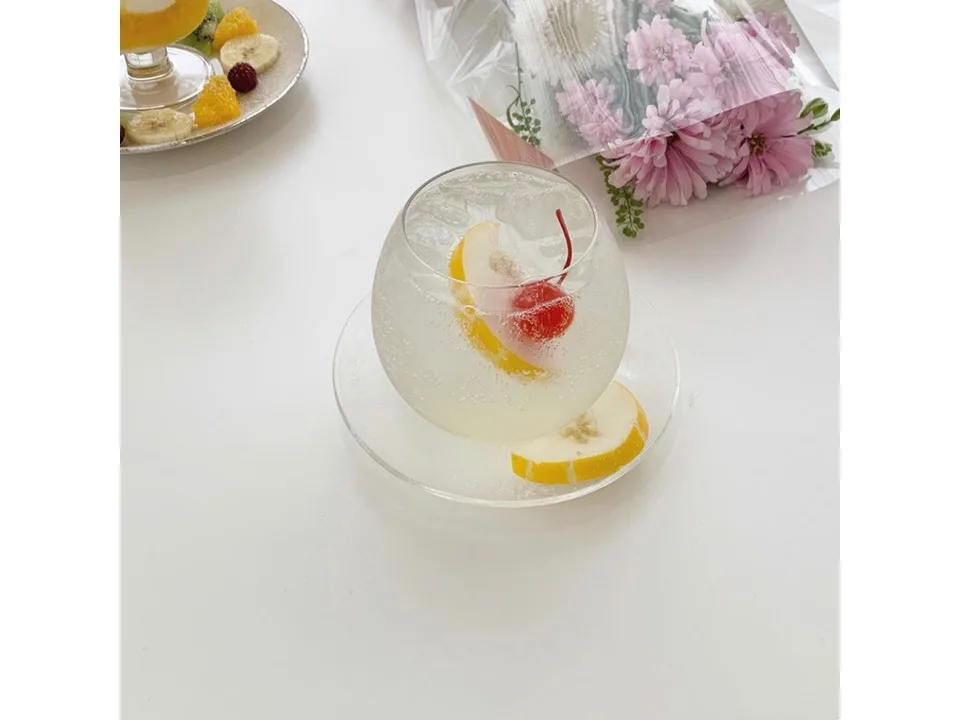 A round glass cup filled with a clear drink, a lemon slice, and a cherry, sitting on a matching glass saucer.
