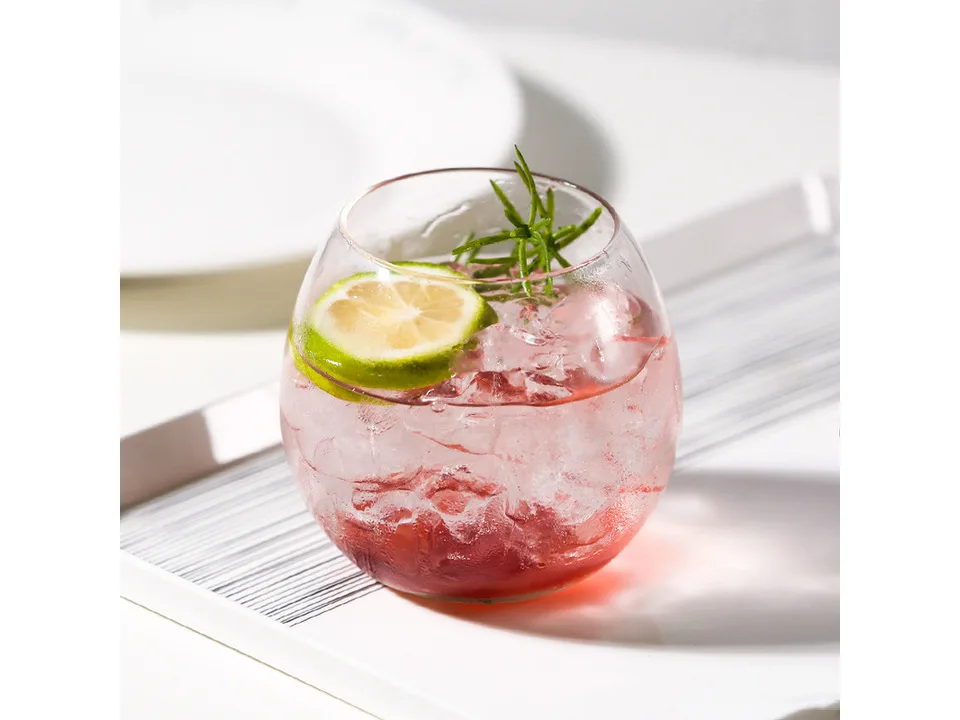 A round pot-belly glass cup filled with a pink drink, ice, a lime slice, and a sprig of rosemary.