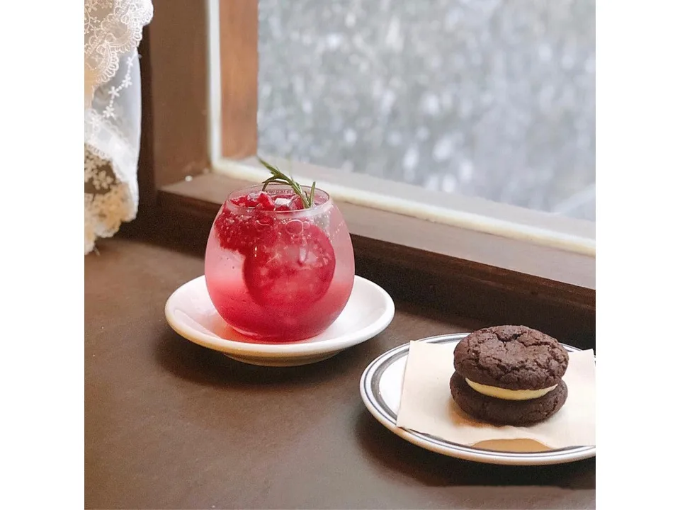 A round glass cup with a red drink and rosemary, placed on a saucer next to a cookie.