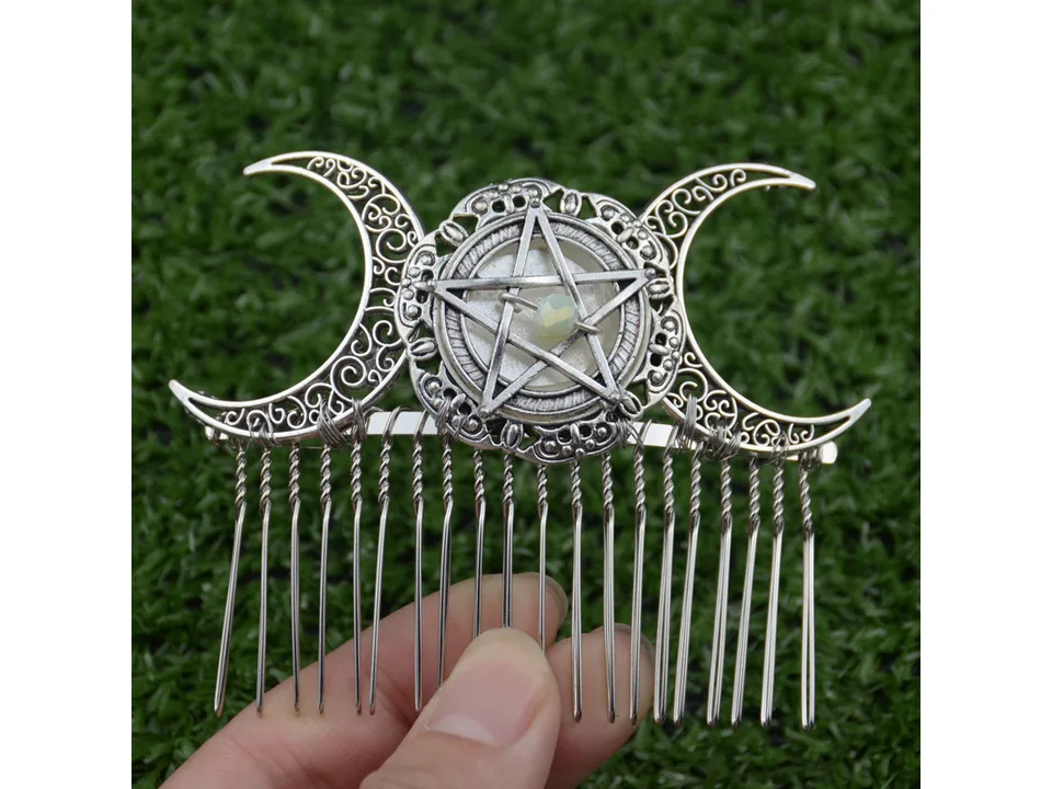 A hand holding a silver pentagram and moon hair comb against a green background.