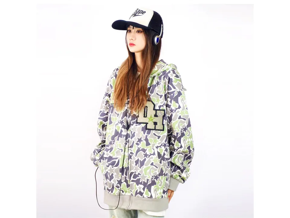Model wearing a green camouflage zip-up hoodie with demon horns on the hood.