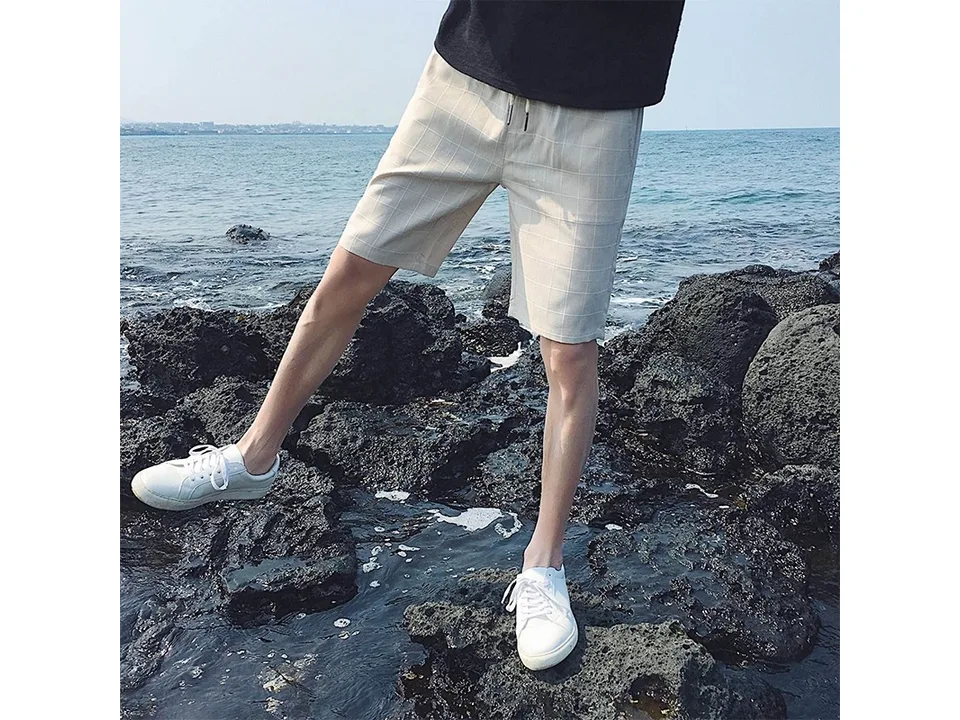 Man wearing beige plaid casual shorts standing on rocks by the sea.