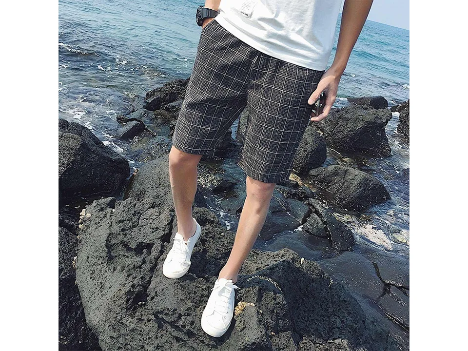Man wearing dark grey plaid casual shorts standing on rocks by the sea.