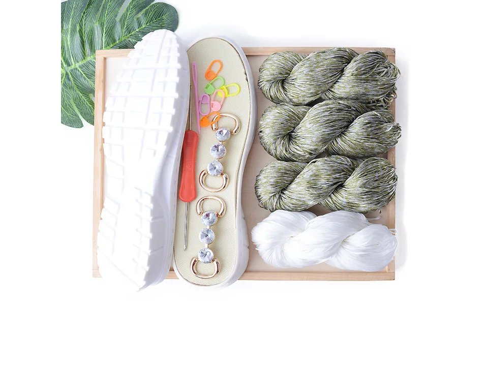 DIY crochet shoe kit with olive green yarn, white yarn, shoe sole, crochet hook, and decorative accessories.
