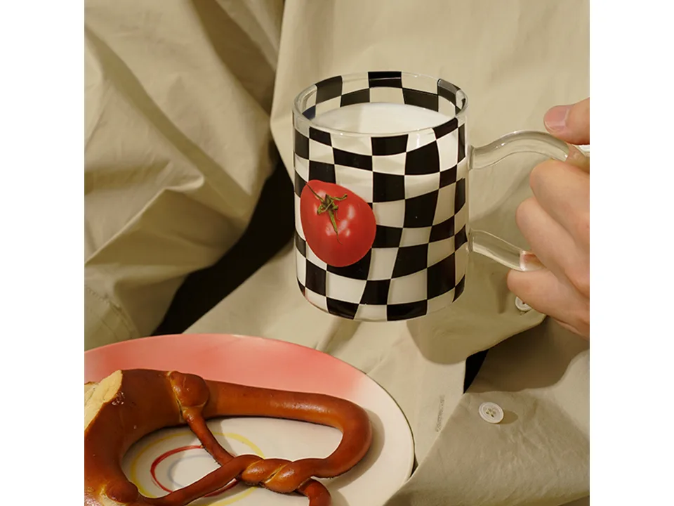 A person holding a checkerboard glass mug with a tomato print next to a plate with a pretzel.