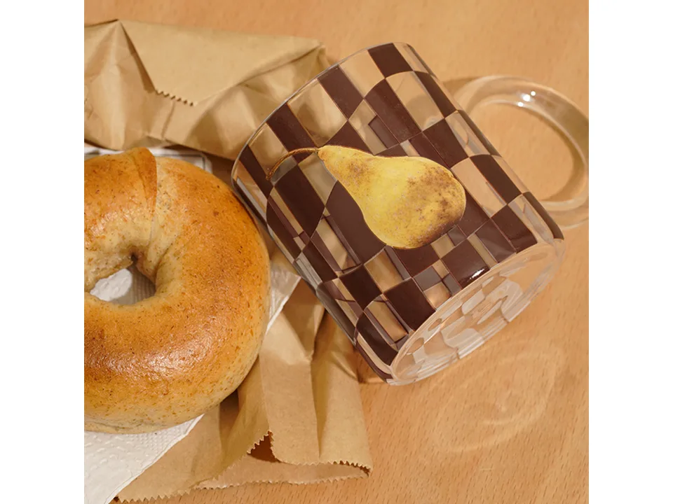 A checkerboard glass mug with a pear print lying on its side next to a bagel.