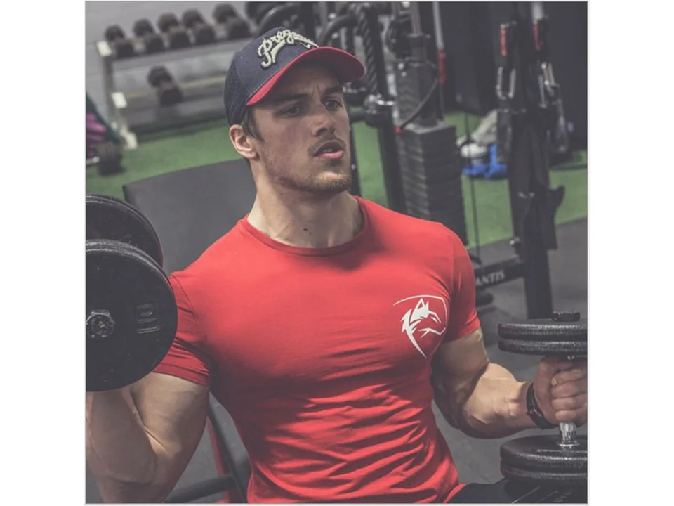 A man lifting dumbbells while wearing a red slim-fit sports t-shirt with a white wolf logo.