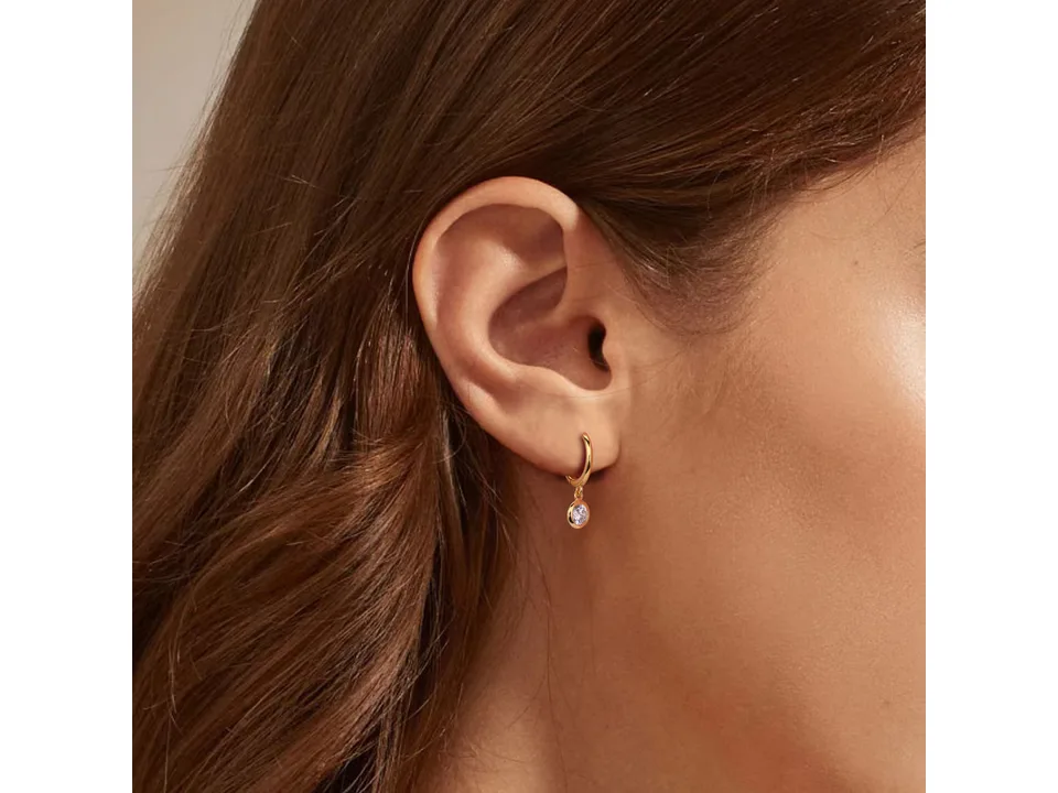 A woman wearing a small gold hoop earring with a dangling pink stone.