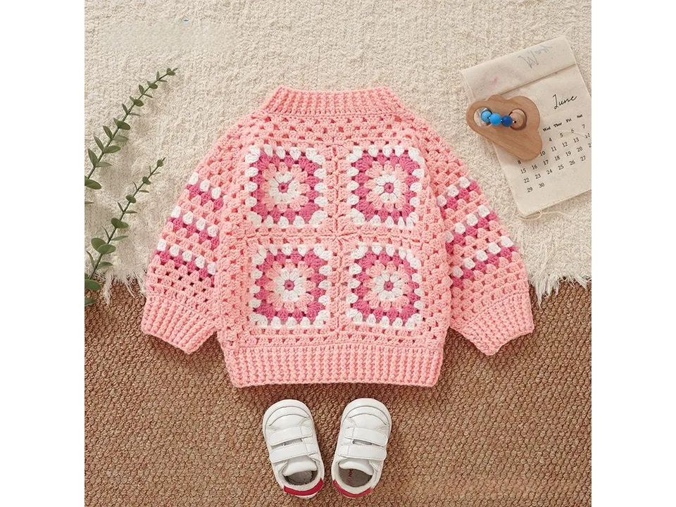 A pink and white crochet-style knitted sweater laid flat with baby shoes.
