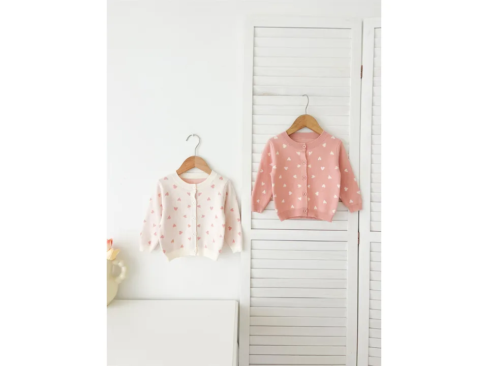 Two baby cardigans, one cream and one pink, both with heart patterns, hanging on wooden hangers.
