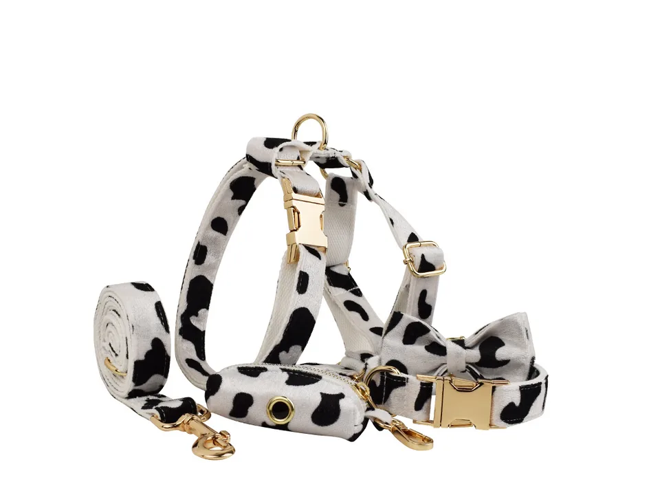 A complete set of cow print dog accessories including a harness, leash, collar, bow tie, and poop bag holder.