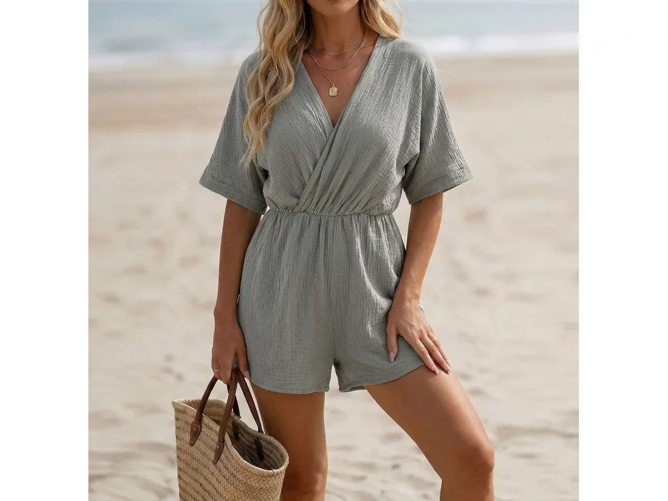 Front view of a woman wearing a sage green V-neck short-sleeve jumpsuit on a beach.