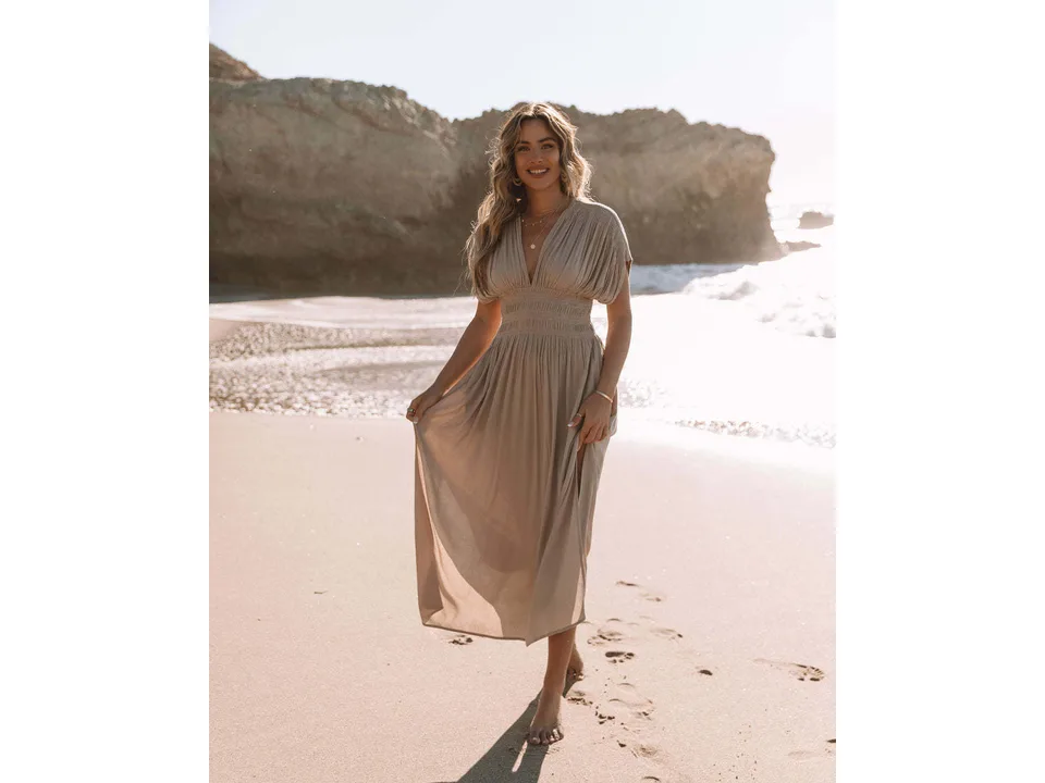 Model walking on the beach wearing a beige maxi dress with a V-neck and cinched waist.