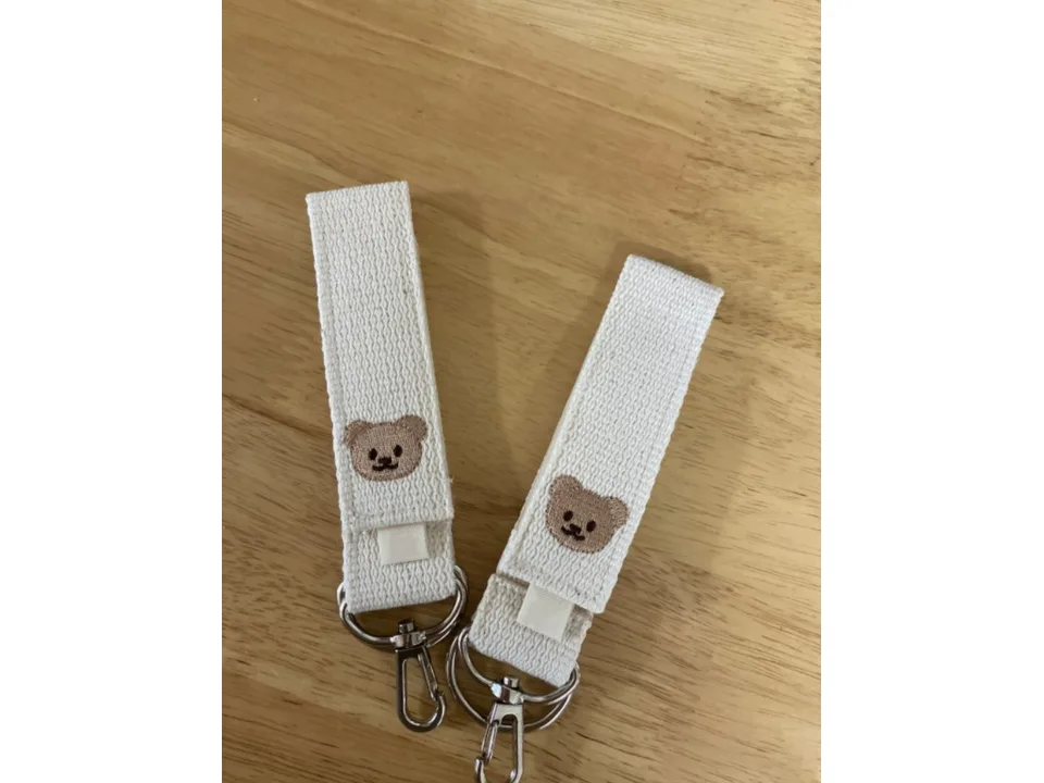Two cream-colored fabric stroller hooks with bear faces lying on a wooden surface.