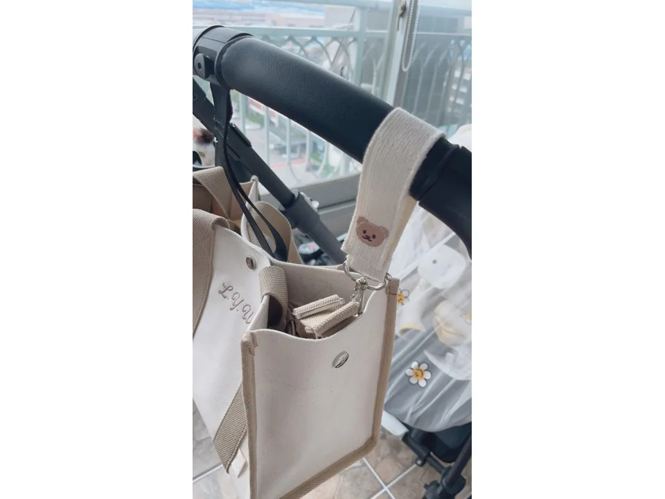 A cream-colored fabric stroller hook with a bear face attached to a stroller handle holding a bag.