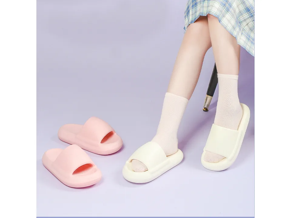 A person wearing white thick-soled slippers with socks, with a pair of pink slippers nearby.
