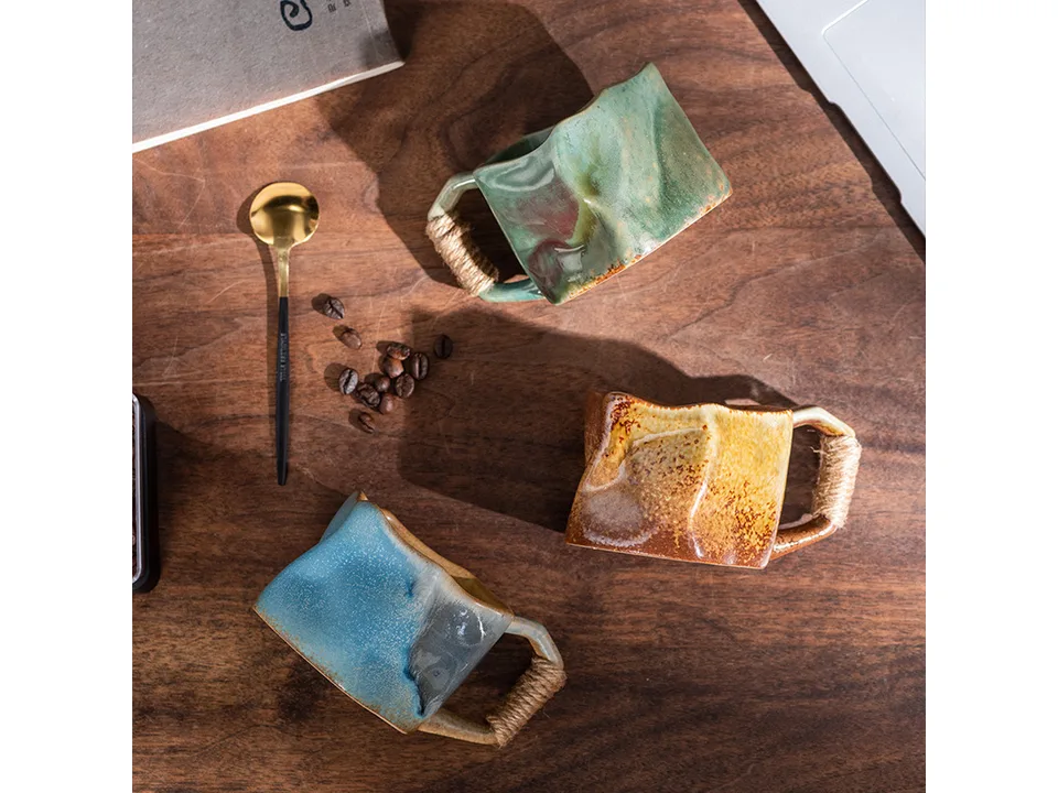 Three different colored irregular ceramic mugs with rope handles arranged on a wooden table.