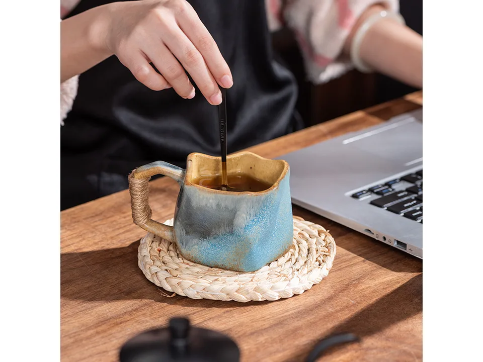 A person stirring a drink in the blue irregular ceramic mug on a wooden desk.