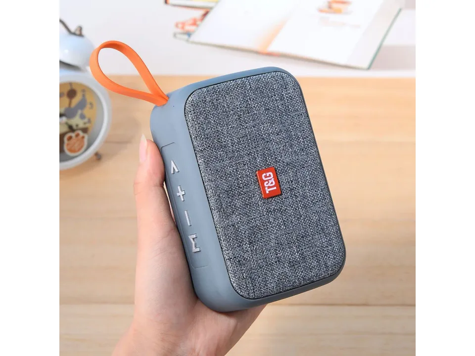 A hand holding a grey portable Bluetooth speaker with an orange carrying loop.