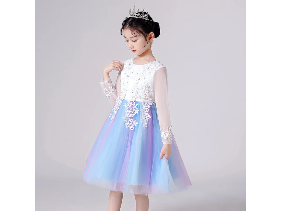 A young girl wearing a white long-sleeved dress with a rainbow gradient mesh skirt and floral lace bodice.