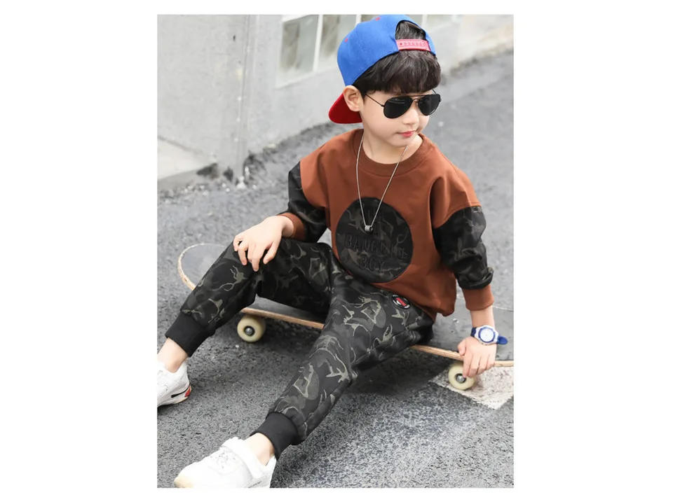 A boy sitting on a skateboard wearing a brown and camouflage two-piece outfit.