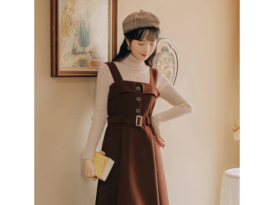 A woman wearing the burgundy pinafore dress over a cream turtleneck sweater.
