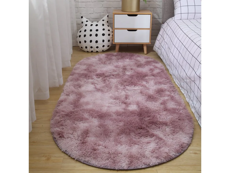 A dusty pink oval shaggy rug placed on a wooden floor next to a bed.
