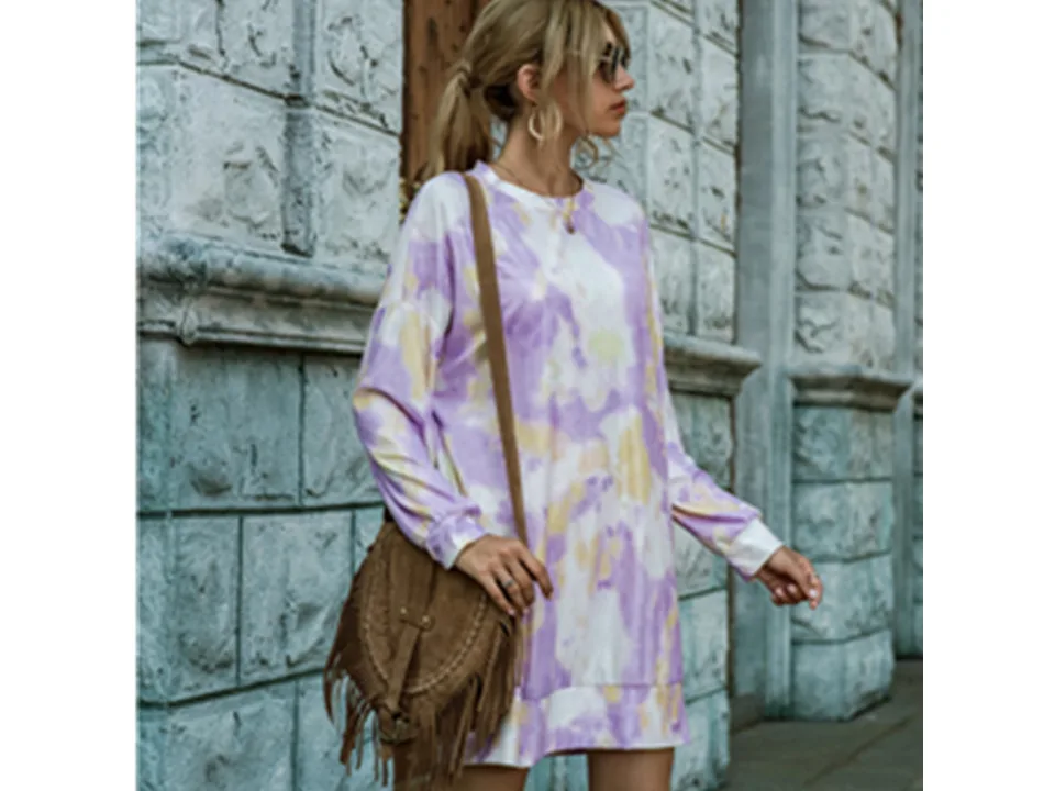 Model wearing a purple and yellow tie-dye long sleeve sweater dress with a brown fringe bag.