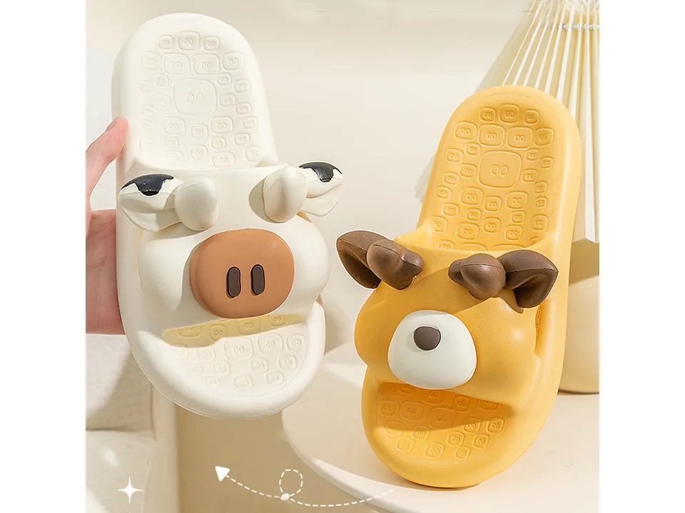 A hand holding a white cow slipper next to a yellow deer slipper.