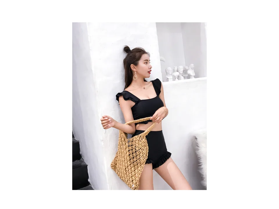 A woman wearing a black high-waisted ruffle bikini set and holding a woven bag.