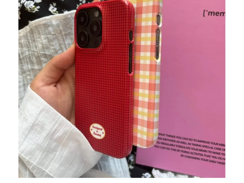 A hand holding a red grid pattern phone case next to a pink plaid phone case.