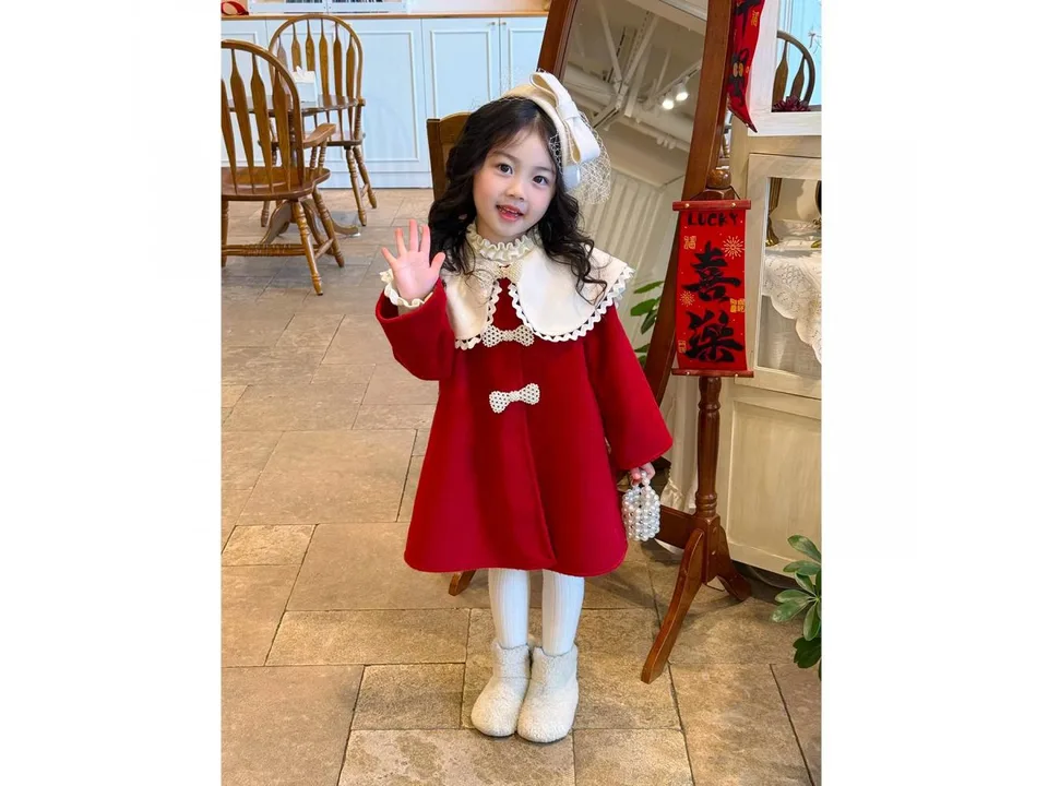 A young girl wearing a red winter coat with a large white collar and pearl bow details, waving.
