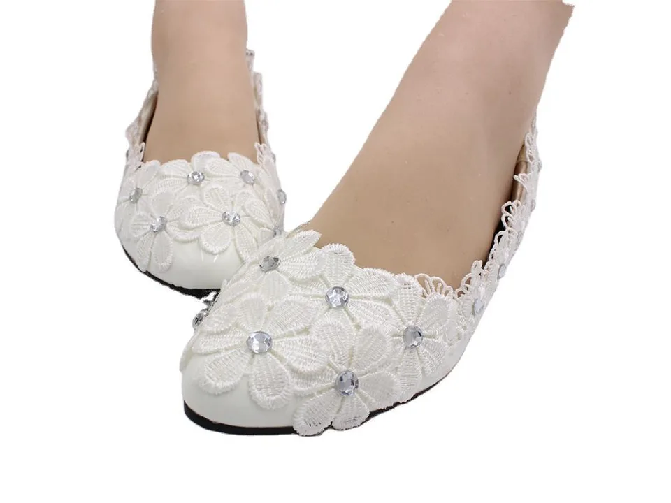 A pair of white lace flat shoes with rhinestone floral embellishments worn on feet.