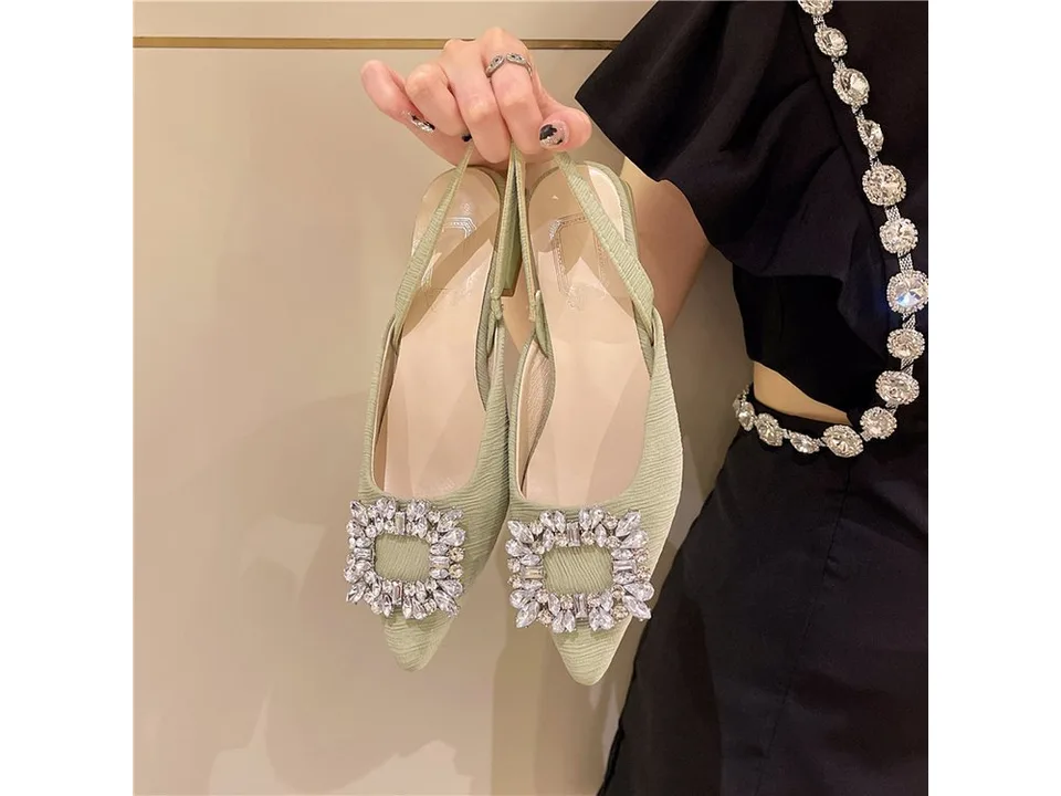 A pair of light green pointed toe slingback heels with a large rhinestone buckle being held by a person.