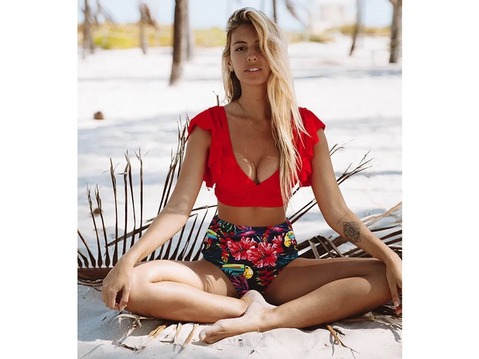 Woman wearing a red ruffle bikini top and floral high-waisted bottoms while sitting on the beach.