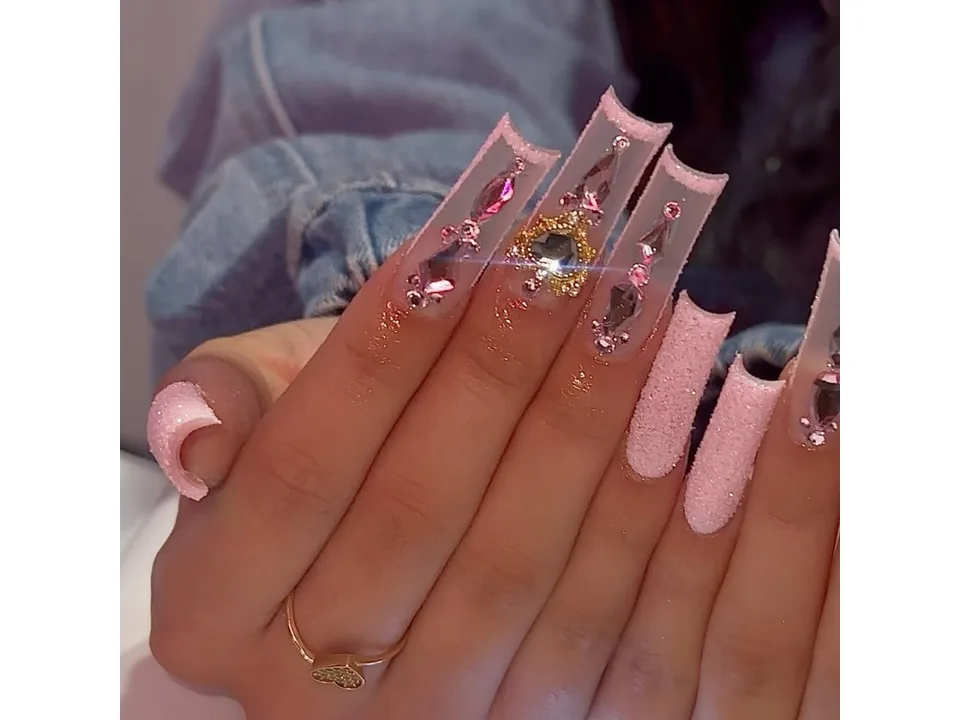 A hand wearing long square press-on nails with pink glitter and rhinestone heart accents.