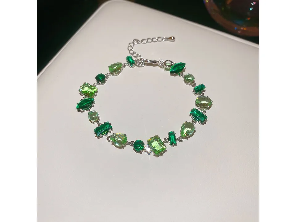 A silver bracelet with various green gemstone shapes on a white surface.