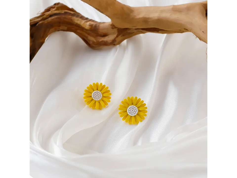 A pair of yellow daisy stud earrings on a white fabric background with a piece of driftwood.