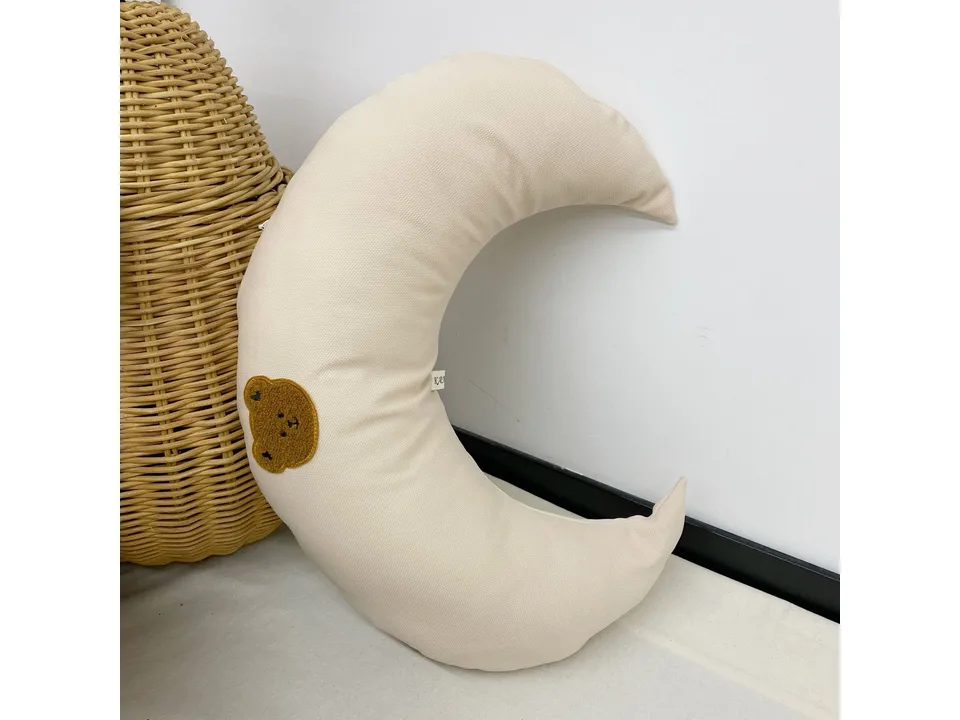 Cream-colored moon-shaped baby pillow with a small brown bear patch.