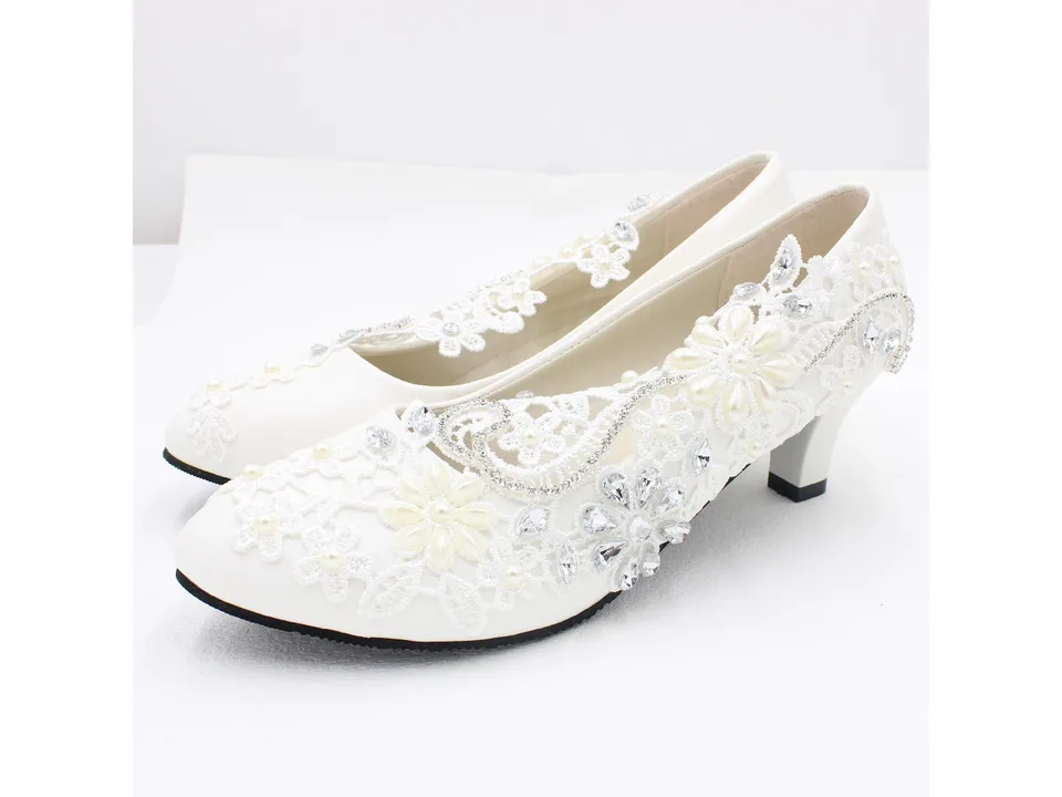 A pair of white lace bridal pumps with floral and rhinestone embellishments.