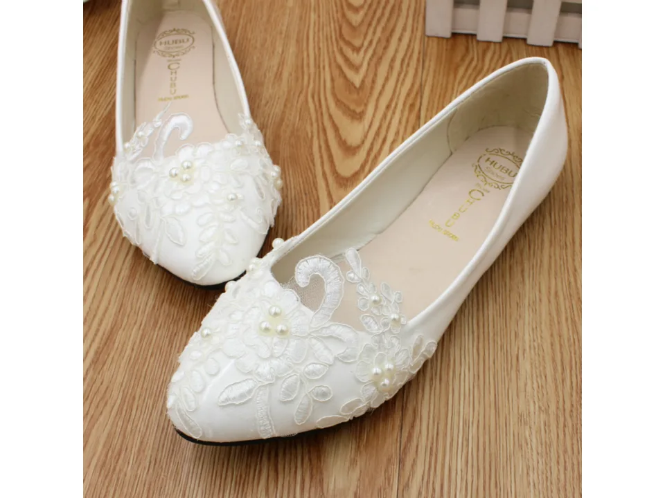 Pair of white lace wedding flats with pearl embellishments on a wooden surface.