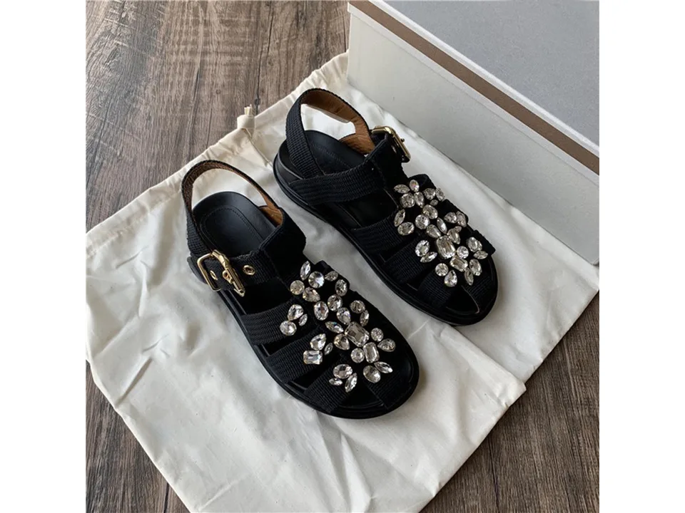 A pair of black closed-toe sandals with rhinestone embellishments on a white dust bag.