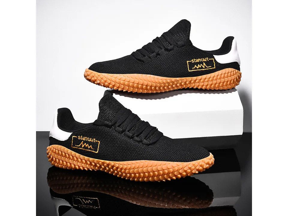 A pair of black low-top sneakers with orange textured soles displayed on a white block.