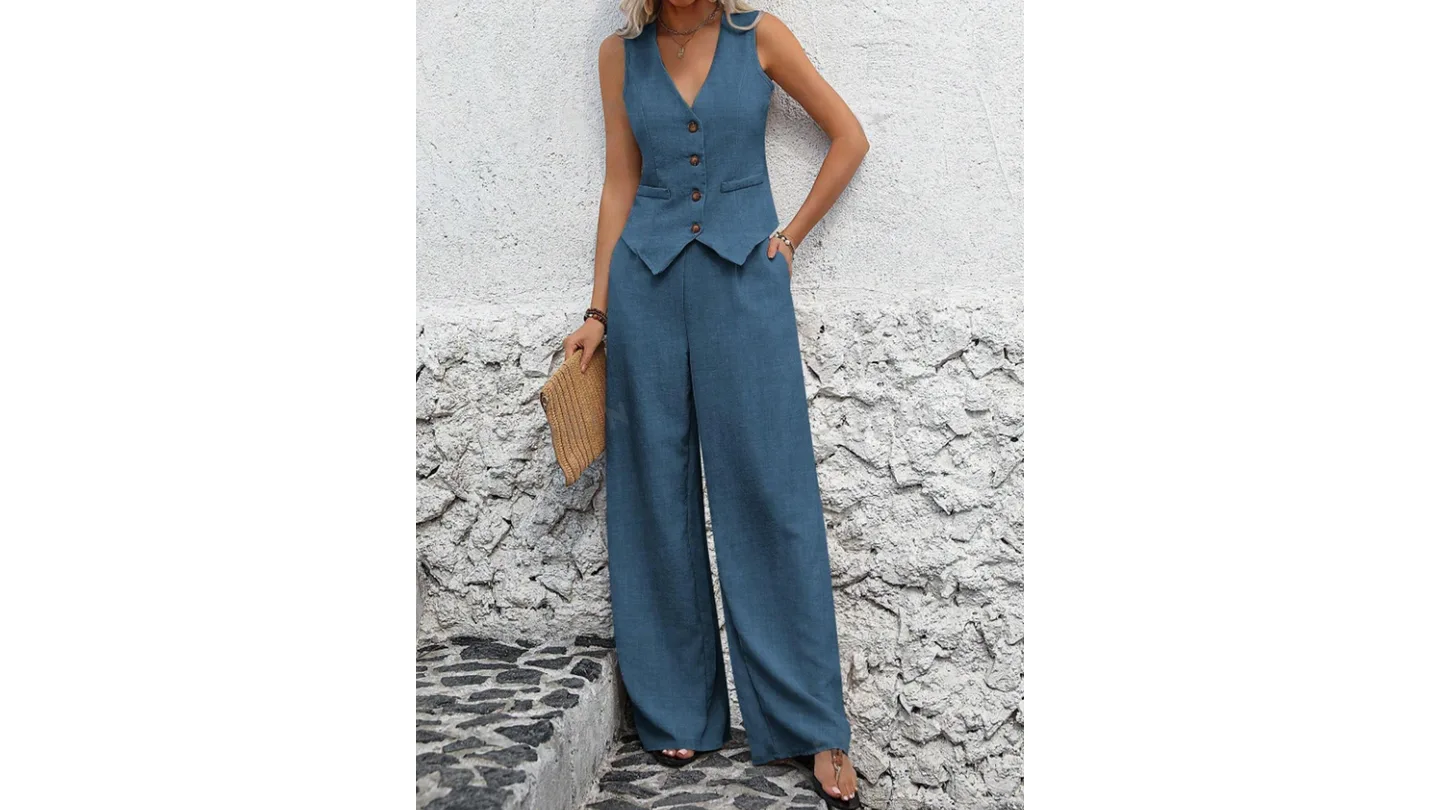 A woman wearing a blue vest and matching wide-leg pants, standing against a textured white wall.